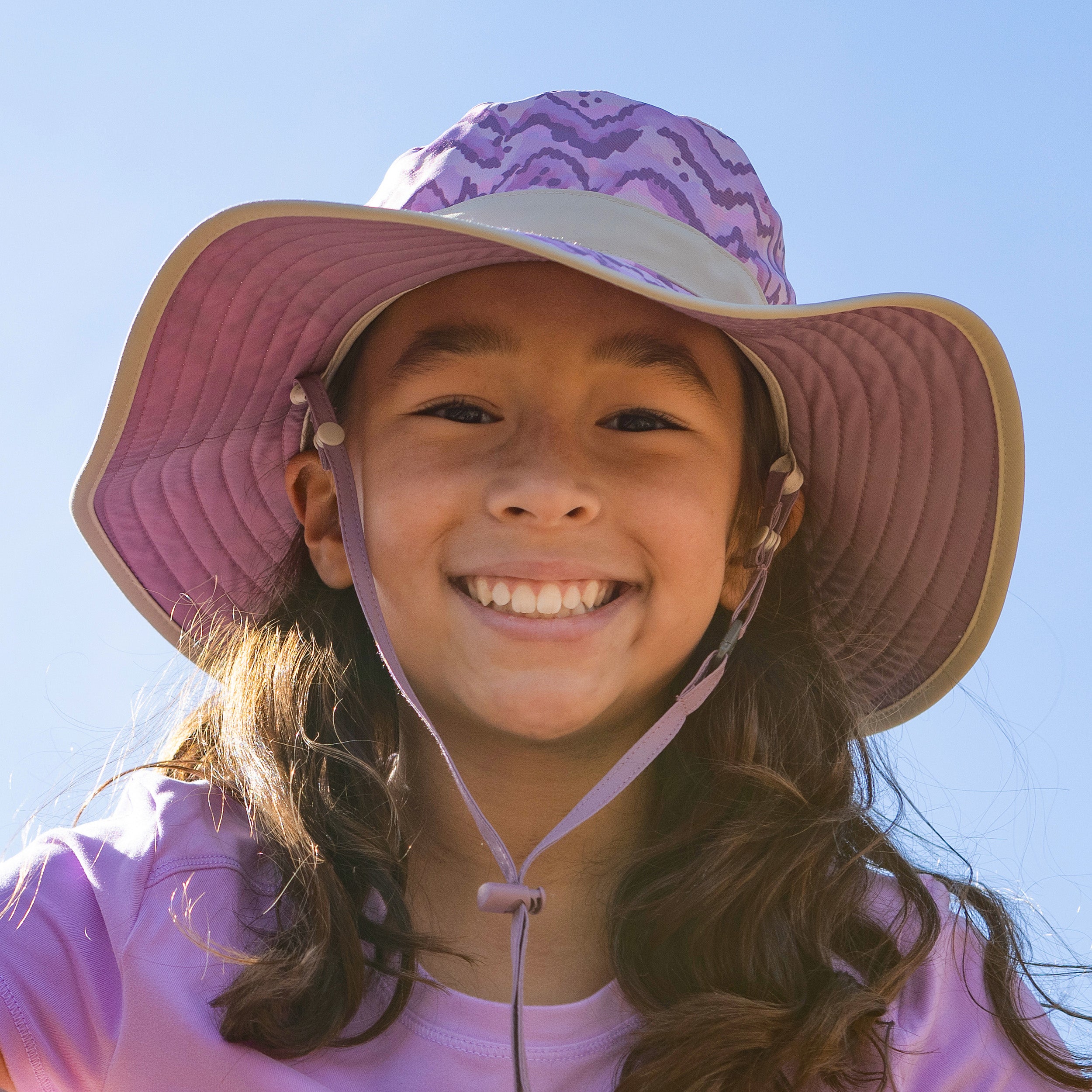 A girl smiling, wearing a Sunday Afternoons Kids' Clear Creek Boonie in the color Wisteria Peaks