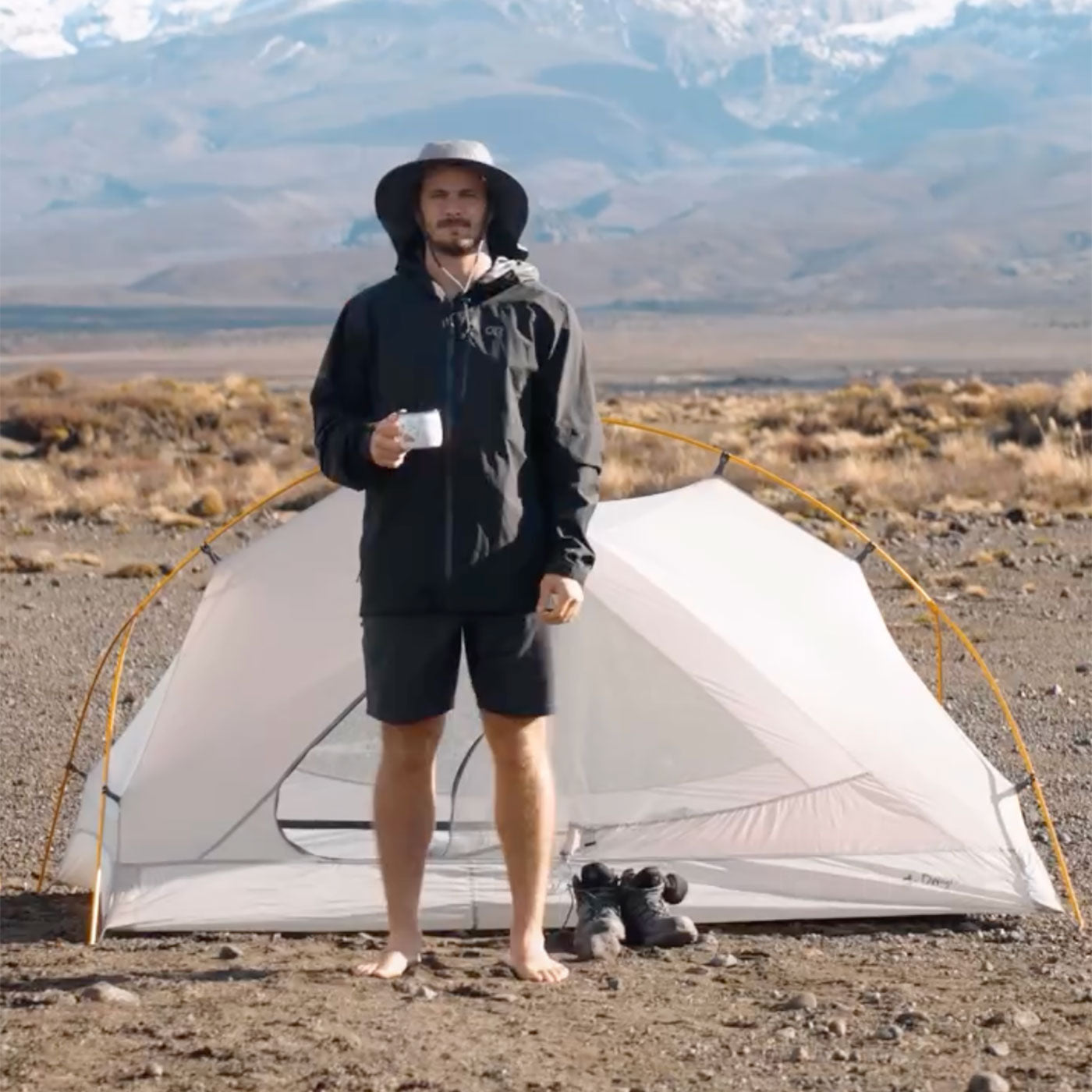 Person standing next to a small tent in a desert landscape with mountains in the background